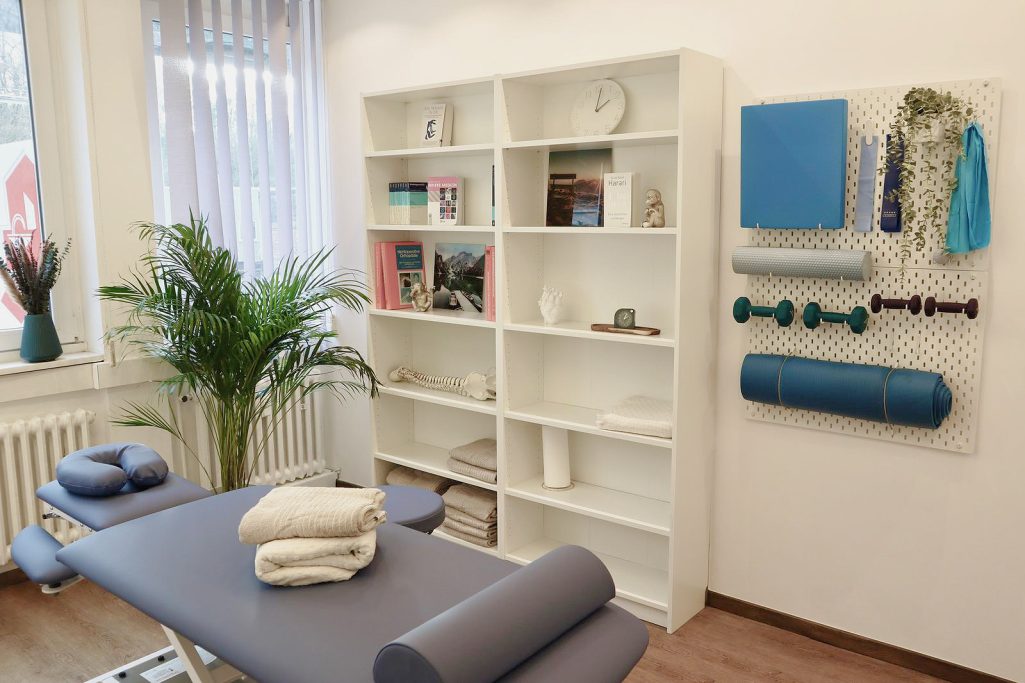 Behandlungsraum 2 Behandlungsraum mit einem Tisch, Regalen und Fitnessgeräten an der Wand.